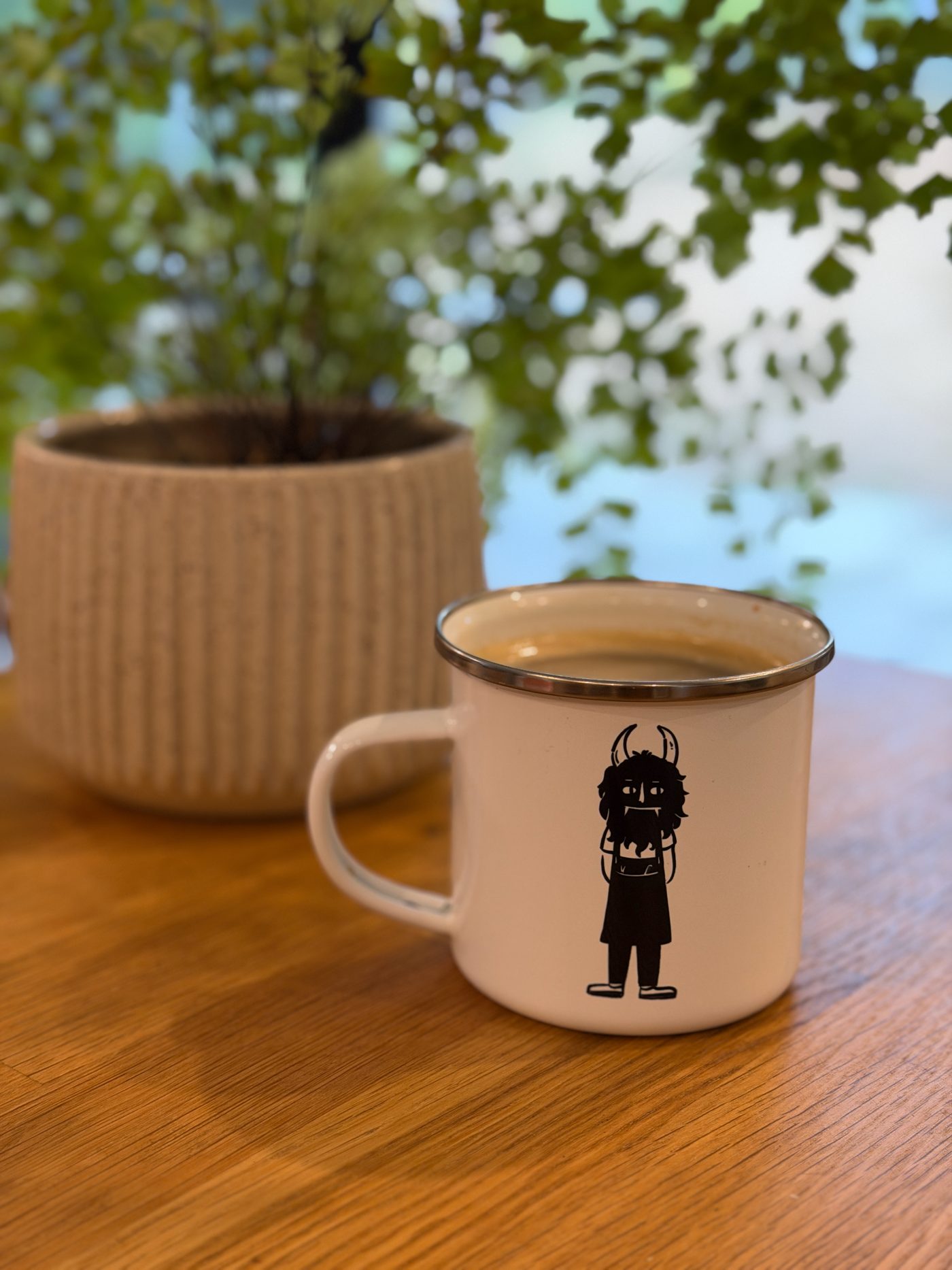 A white enamel mug of coffee printed with the salonMonster monster, on a wooden table beside a potted plant