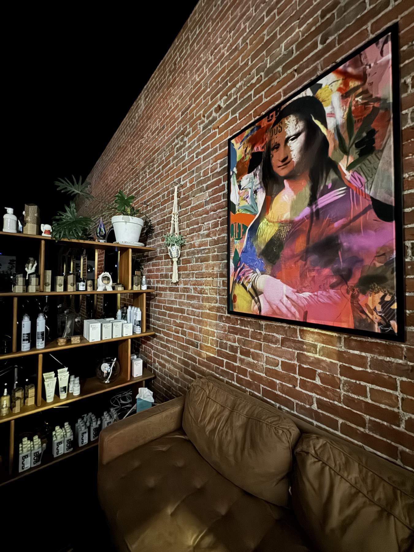 A leather sofa under a colourful painting on a brick wall, beside shelves of salon products
