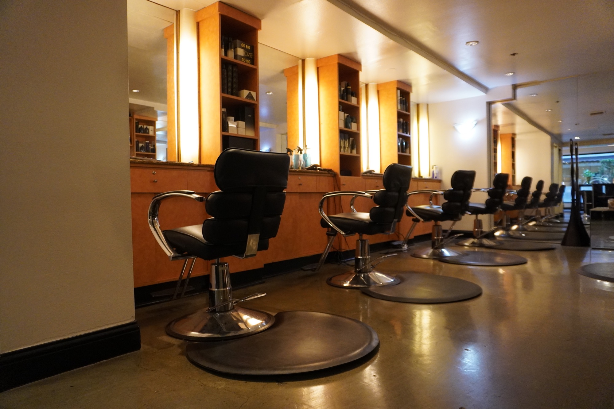 A row of black styling chairs along a wall of lit mirrors in a salon